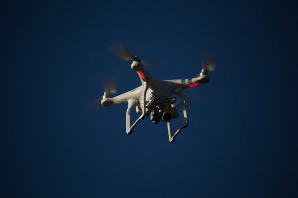 A white drone with four rotors is flying in the sky. The drone is visible against a dark blue sky and appears to have a camera mounted at its base. The rotors are in motion, and there are red lights on the drone's body.