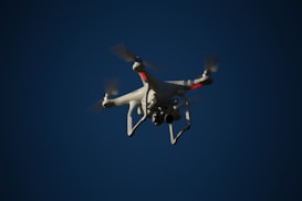A white drone with four rotors is flying in the sky. The drone is visible against a dark blue sky and appears to have a camera mounted at its base. The rotors are in motion, and there are red lights on the drone's body.