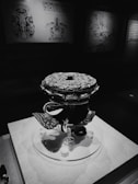 A dark, intricately designed artifact with a patterned surface, displayed on a pedestal in a dimly lit exhibition. Background features maps and diagrams with faded illustrations.