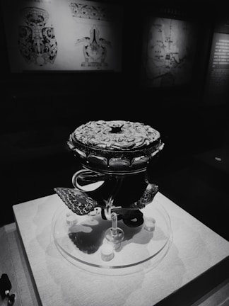 A dark, intricately designed artifact with a patterned surface, displayed on a pedestal in a dimly lit exhibition. Background features maps and diagrams with faded illustrations.