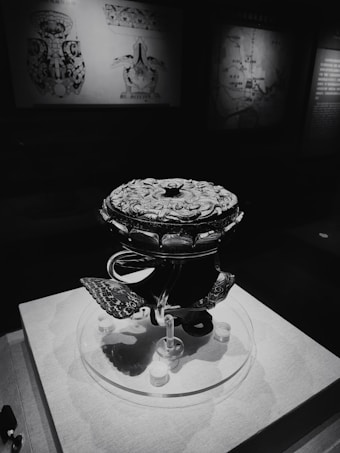 A dark, intricately designed artifact with a patterned surface, displayed on a pedestal in a dimly lit exhibition. Background features maps and diagrams with faded illustrations.