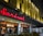A modern building facade featuring a prominent bright red sign with the name 'Sarinah'. Beneath it is an illuminated window display with the word 'Dewata', and interior lights and decor visible inside. The exterior design includes unique golden architectural panels.