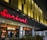 A modern building facade featuring a prominent bright red sign with the name 'Sarinah'. Beneath it is an illuminated window display with the word 'Dewata', and interior lights and decor visible inside. The exterior design includes unique golden architectural panels.
