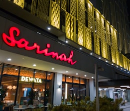 A modern building facade featuring a prominent bright red sign with the name 'Sarinah'. Beneath it is an illuminated window display with the word 'Dewata', and interior lights and decor visible inside. The exterior design includes unique golden architectural panels.