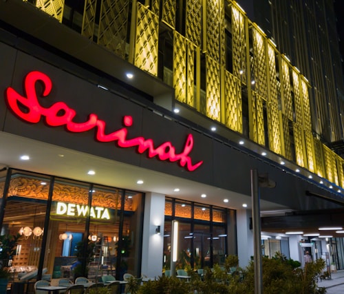 A modern building facade featuring a prominent bright red sign with the name 'Sarinah'. Beneath it is an illuminated window display with the word 'Dewata', and interior lights and decor visible inside. The exterior design includes unique golden architectural panels.