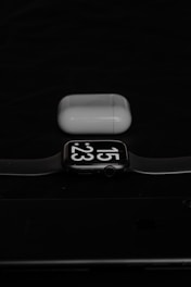 A smartwatch displaying the time 15:23 is placed in front of a pair of wireless earphones in their charging case. The background is dark, creating a contrasting effect with the white and black elements of the watch and earphones.