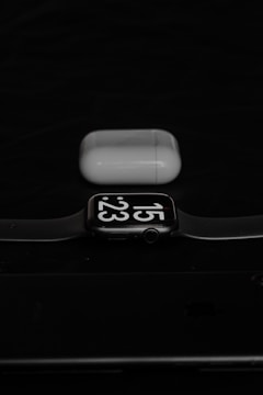 A smartwatch displaying the time 15:23 is placed in front of a pair of wireless earphones in their charging case. The background is dark, creating a contrasting effect with the white and black elements of the watch and earphones.
