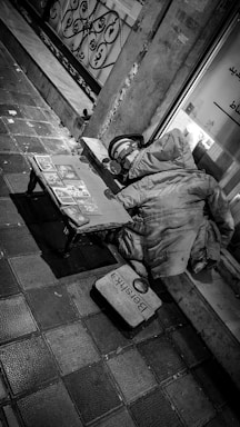 A black and white photograph features an urban street scene. On the left, a small wooden table or bench holds various spread-out items, possibly books or cards. Adjacent to it, a bulky, worn-out coat is laid on the ground, suggesting the presence of a homeless person. Nearby, a helmet and a rectangular sign, which appears to be in front of a shop window with some text, are visible. The pavement is made of tiles and appears somewhat grimy, enhancing the gritty urban feel.