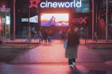 A group of people are walking towards an illuminated cinema entrance at night. The signage displays 'cineworld' prominently above the entrance, with additional signs for 'SuperScreen' and '4DX' visible. A large digital screen above the entrance shows a colorful image.