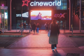 A group of people are walking towards an illuminated cinema entrance at night. The signage displays 'cineworld' prominently above the entrance, with additional signs for 'SuperScreen' and '4DX' visible. A large digital screen above the entrance shows a colorful image.
