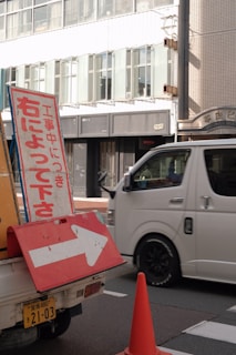 A street scene featuring a traffic sign with Japanese text and a right-pointing arrow, situated on a vehicle. A white van is passing by on the road, and an orange traffic cone is nearby. In the background, there is a modern building with large windows.