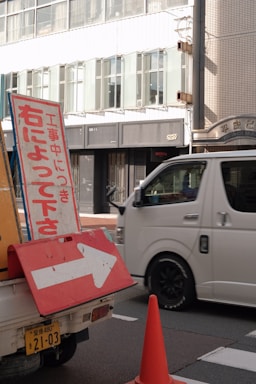 A street scene featuring a traffic sign with Japanese text and a right-pointing arrow, situated on a vehicle. A white van is passing by on the road, and an orange traffic cone is nearby. In the background, there is a modern building with large windows.