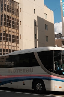 A large bus with the name 'IYOTETSU' is parked on a street beside a tall building. The building has a grid of windows and a shadow cast on its facade. Part of a sign with Japanese characters is visible in the background.