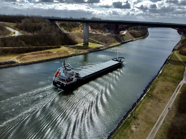A long cargo ship travels along a wide canal, creating waves in the water. A bridge spans the canal, with grassy banks and sparse trees lining the sides. The sky above is filled with scattered clouds.