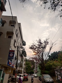 A street scene at sunset featuring a multi-story building with a dental clinic sign. The street is lined with trees, one of which has pink flowers and partially obscures the sun. Cars and motorbikes are parked along the sides, and a person walks down the street wearing purple clothing.