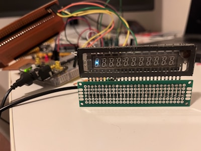 A setup involving electronic components with wires is visible, featuring a digital display and a circuit board with multiple connectors. Various colored wires are connected to the components, and a power cable is plugged in. The arrangement appears to be part of a prototype or a DIY electronics project.