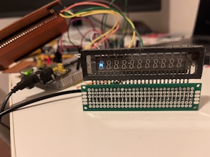 A setup involving electronic components with wires is visible, featuring a digital display and a circuit board with multiple connectors. Various colored wires are connected to the components, and a power cable is plugged in. The arrangement appears to be part of a prototype or a DIY electronics project.