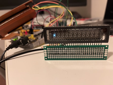 A setup involving electronic components with wires is visible, featuring a digital display and a circuit board with multiple connectors. Various colored wires are connected to the components, and a power cable is plugged in. The arrangement appears to be part of a prototype or a DIY electronics project.