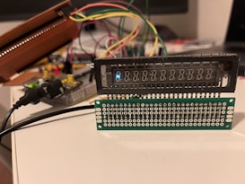 A setup involving electronic components with wires is visible, featuring a digital display and a circuit board with multiple connectors. Various colored wires are connected to the components, and a power cable is plugged in. The arrangement appears to be part of a prototype or a DIY electronics project.