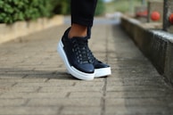 A pair of black sneakers with white soles is being worn by a person standing on a stone-paved path. The background includes a low stone wall and red railings, with greenery visible on one side, creating a casual and outdoor setting.