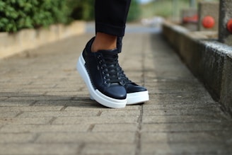 A pair of black sneakers with white soles is being worn by a person standing on a stone-paved path. The background includes a low stone wall and red railings, with greenery visible on one side, creating a casual and outdoor setting.