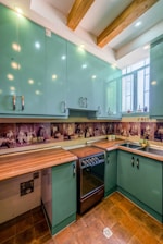 A kitchen with teal cabinets and wooden countertops. The backsplash features artistic sepia-toned images. The room has a modern stove and a stainless steel sink under a window. Exposed wooden beams are visible on the ceiling, with recessed lighting.
