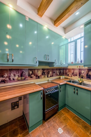 A kitchen with teal cabinets and wooden countertops. The backsplash features artistic sepia-toned images. The room has a modern stove and a stainless steel sink under a window. Exposed wooden beams are visible on the ceiling, with recessed lighting.