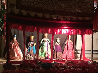 A display of five elaborately dressed puppets inside a decorative enclosure. The setting is dimly lit with red and warm tones, and each puppet wears traditional attire with intricate patterns and vibrant colors. Above the puppets, a richly adorned fabric covers the top of the display.