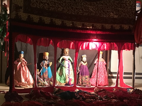 A display of five elaborately dressed puppets inside a decorative enclosure. The setting is dimly lit with red and warm tones, and each puppet wears traditional attire with intricate patterns and vibrant colors. Above the puppets, a richly adorned fabric covers the top of the display.