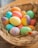 A basket filled with brightly colored Easter eggs rests on a bed of straw. The eggs display a variety of colors, including blue, pink, orange, yellow, and multicolored patterns. A small, blurred bird decoration appears on the edge of the basket.