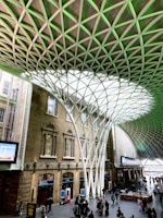 An architectural interior with a large open space featuring an impressive geometric roof structure made from interlocking metal beams. Sunlight filters through the translucent panels, casting patterns on the floor. Below, people are gathered, some walking and others standing in small groups. The surrounding structure includes brick walls with arched windows.