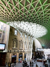 An architectural interior with a large open space featuring an impressive geometric roof structure made from interlocking metal beams. Sunlight filters through the translucent panels, casting patterns on the floor. Below, people are gathered, some walking and others standing in small groups. The surrounding structure includes brick walls with arched windows.