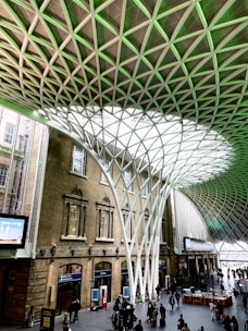 An architectural interior with a large open space featuring an impressive geometric roof structure made from interlocking metal beams. Sunlight filters through the translucent panels, casting patterns on the floor. Below, people are gathered, some walking and others standing in small groups. The surrounding structure includes brick walls with arched windows.