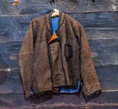 A brown wool jacket hangs on a rustic wooden wall. The jacket features black trim and a simple design with a single visible pocket. It is partially open, revealing a hint of blue lining and an orange interior. The wooden wall behind has a rugged, weathered texture with visible knots and grain patterns.