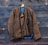 A brown wool jacket hangs on a rustic wooden wall. The jacket features black trim and a simple design with a single visible pocket. It is partially open, revealing a hint of blue lining and an orange interior. The wooden wall behind has a rugged, weathered texture with visible knots and grain patterns.