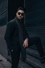 A stylish man wearing sunglasses and a patterned scarf poses with one leg on a step, set against a dark urban backdrop. He has a well-groomed beard and wears a watch on his left wrist. The setting conveys a sense of modernity and elegance.
