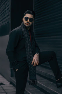 Editorial photo of a male model wearing a beige tailored suit with a playful patterned scarf, standing in front of a modern art installation