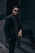 A stylish man wearing sunglasses and a patterned scarf poses with one leg on a step, set against a dark urban backdrop. He has a well-groomed beard and wears a watch on his left wrist. The setting conveys a sense of modernity and elegance.