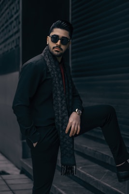 A stylish man wearing sunglasses and a patterned scarf poses with one leg on a step, set against a dark urban backdrop. He has a well-groomed beard and wears a watch on his left wrist. The setting conveys a sense of modernity and elegance.