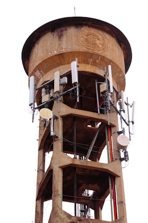 A tall, cylindrical water tower structure made of concrete with an aged and weathered appearance. The upper section has intricate designs embossed in the concrete, possibly indicating a historical or official significance. Several telecommunication antennas and equipment are attached around the upper portion, suggesting dual use for water storage and communication. The structure has metal beams and reinforcements visible, adding to its industrial appearance.