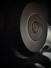 Close-up view of a headphone earcup with a prominent logo on it. The lighting creates a dramatic shadow effect, highlighting the curved surface and metallic texture.
