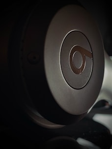 Close-up view of a headphone earcup with a prominent logo on it. The lighting creates a dramatic shadow effect, highlighting the curved surface and metallic texture.