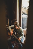 A person with medium-length hair plays an acoustic guitar while seated in a chair near a glass door. The scene is softly lit, suggesting a relaxed, introspective mood. The person wears a long-sleeve shirt and jeans, and there are chairs and a foosball table visible in the background.