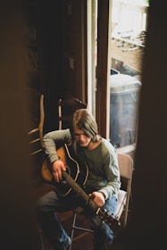 A person with medium-length hair plays an acoustic guitar while seated in a chair near a glass door. The scene is softly lit, suggesting a relaxed, introspective mood. The person wears a long-sleeve shirt and jeans, and there are chairs and a foosball table visible in the background.