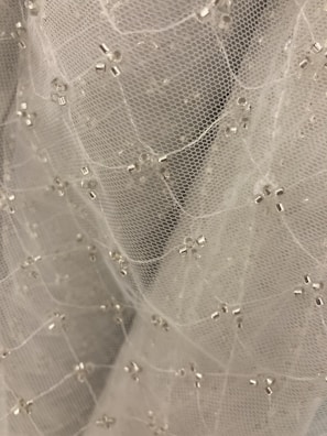 A close-up view of delicate white mesh fabric adorned with small, shiny, silver beads arranged in a subtle pattern. The fabric appears soft and lightweight, creating a sense of elegance and intricacy.
