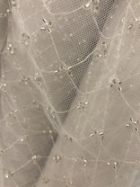 A close-up view of delicate white mesh fabric adorned with small, shiny, silver beads arranged in a subtle pattern. The fabric appears soft and lightweight, creating a sense of elegance and intricacy.