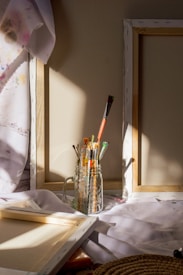 A collection of paintbrushes is neatly arranged in a transparent glass jar. The jar is placed on a white cloth, which partially covers empty wooden canvas frames. Sunlight streams across the scene, casting soft shadows and illuminating the brushes and cloth. Splashes of color are visible on a fabric beside the frames.