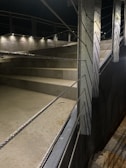 A well-lit outdoor staircase with concrete steps, metal railings, and multiple spotlights illuminating the area. The railings have horizontal metal cables and the surroundings appear clean and modern.