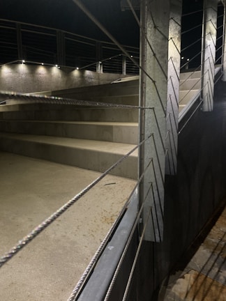 A well-lit outdoor staircase with concrete steps, metal railings, and multiple spotlights illuminating the area. The railings have horizontal metal cables and the surroundings appear clean and modern.