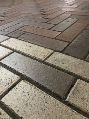 A close-up view of a brick pavement with a herringbone pattern. The bricks vary in color, including shades of gray, beige, and brown, creating a textured and ordered appearance.
